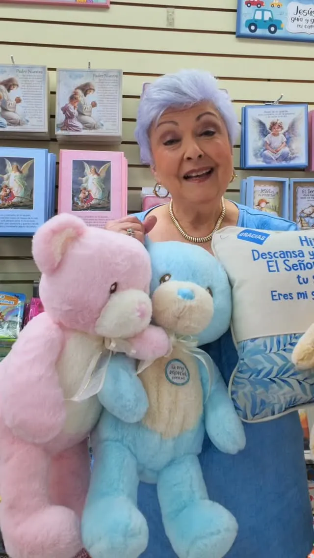 Osos de peluche rosado y celeste y un cojín con versículo, frente a cuadros de ángeles en una tienda Bisel